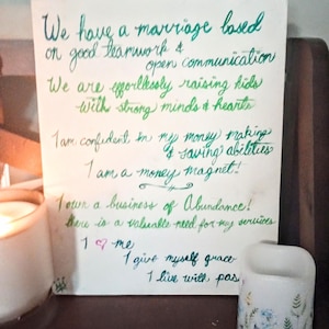 Op de afbeelding: Een handgeschreven notitie op een witte achtergrond met groene en blauwe inkt. De notitie luidt: "We hebben een huwelijk dat is gebaseerd op goed teamwork en open communicatie. We brengen moeiteloos kinderen groot met sterke geesten en harten. Ik ben vol vertrouwen in mijn geldverdienende en spaarcapaciteiten. Ik ben een geldmagneet! Ik bezit een bedrijf van overvloed! Er is een waardevolle behoefte aan mijn diensten. Ik hou van mezelf. Ik geef mezelf genade. Ik leef met passie."