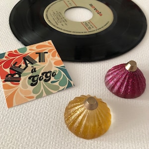 Può includere: Due tappi per bottiglia decorativi, uno dorato e uno rosa, con una parte superiore dorata. I tappi hanno una finitura testurizzata e scintillante. Un disco in vinile vintage e una carta con il testo "Beat à Go Go" sono sullo sfondo.