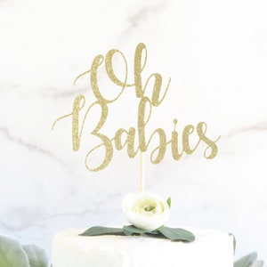 Op de afbeelding: Een gouden glittertaarttopper met de tekst "Oh Babies" op een witte taart met een witte bloem erop.