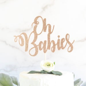 May include: A gold glitter cake topper with the text "Oh Babies" on a white cake with a white flower.