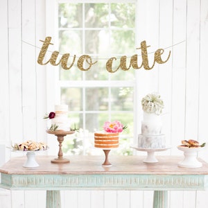 May include: A light blue table with a wooden top, decorated with a gold glitter banner that reads "two cute". There are four cake stands with cakes on them, two with white frosting and two with naked cake.