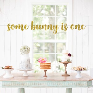 May include: Gold glitter banner with the text "some bunny is one". The banner is hanging above a table with three cakes on cake stands. The cakes are decorated with flowers and greenery.