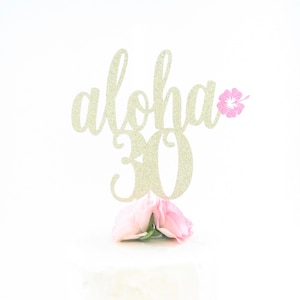 May include: A white cake with a gold glitter "Aloha 30" cake topper and pink flowers.