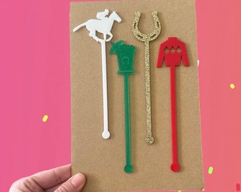 May include: Four cocktail stirrers with horse racing designs. The stirrers are white, green, gold glitter, and red. The designs include a jockey on a horse, a horseshoe, and racing silks. The stirrers are displayed on a brown card.