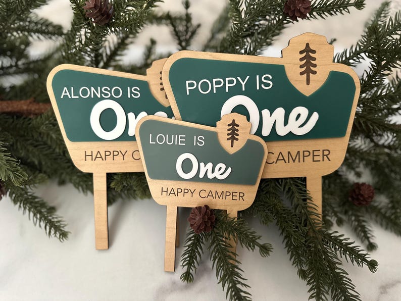 May include: Wooden signposts with green and white text that read "Alonso is One Happy Camper", "Poppy is One Happy Camper", and "Louie is One Happy Camper". Each sign has a tree graphic.