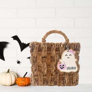 May include: A brown woven basket with a handle, decorated with a pink and white ghost cutout with the text "Valerie" and a pumpkin. The basket is on a white brick background with black bats and a spider.
