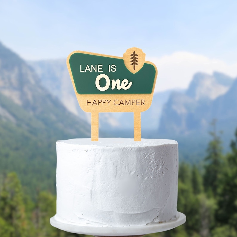 May include: A wooden cake topper with a green and tan sign that reads "LANE IS ONE HAPPY CAMPER." The sign has a tree graphic and is placed on top of a white frosted cake. The background is a blurred image of mountains and trees.