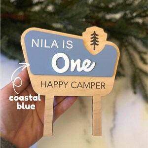 May include: Wooden sign with a coastal blue and natural wood color scheme. The sign reads "NILA IS One HAPPY CAMPER" in white and black lettering. A tree graphic is on the top right corner. The sign has two posts at the bottom.