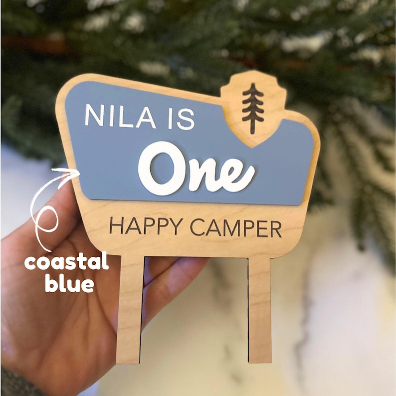 May include: Wooden sign with a coastal blue and natural wood color scheme. The sign reads "NILA IS One HAPPY CAMPER" in white and black lettering. A tree graphic is on the top right corner. The sign has two posts at the bottom.