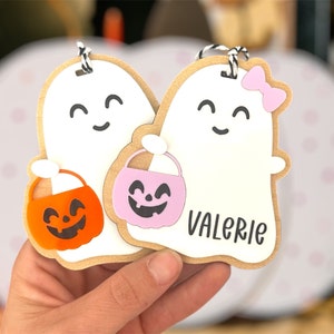 May include: Two white ghost-shaped Halloween treat bags with smiling faces. One ghost has a pink bow and a pink pumpkin bucket. The other ghost has an orange pumpkin bucket. The bag with the pink bow has the name "Valerie" written on it.