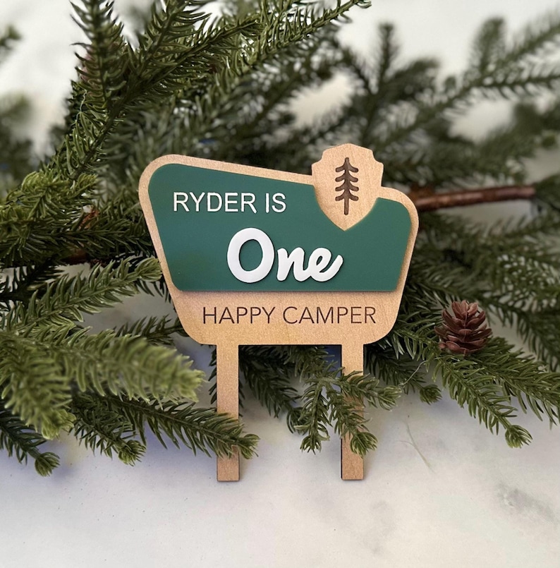May include: A wooden sign with the text "Ryder is One Happy Camper." The sign has a green and tan color scheme with a tree graphic. The sign is designed to look like a park sign.