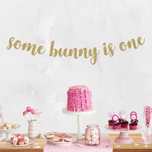 May include: Gold glitter banner with the text "some bunny is one" hanging above a pink and white dessert table with cupcakes, cake pops, and a pink cake.