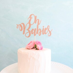 May include: A white cake with pink flowers and a rose gold glitter cake topper that says "Oh Babies".