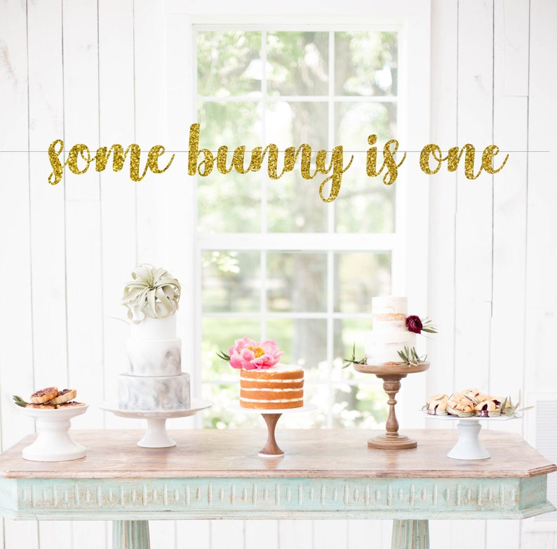 May include: Gold glitter banner with the text "some bunny is one". The banner is hanging above a table with three cakes on cake stands. The cakes are decorated with flowers and greenery.