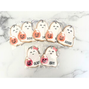 May include: A set of eight white ghost-shaped ornaments with black eyes and mouths. Each ghost is holding a small orange pumpkin bucket and has a name written on it: Ashton, Olivia, Aria, Rehan, Liv, R, and two others. The ornaments are packaged in clear plastic bags.