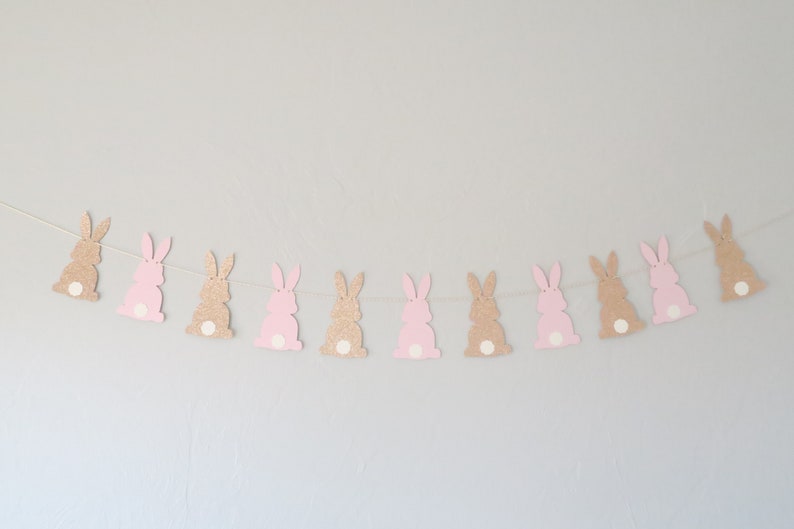May include: A garland of pink and brown glitter bunny cutouts strung on a white string. The bunnies have white spots on their tails.