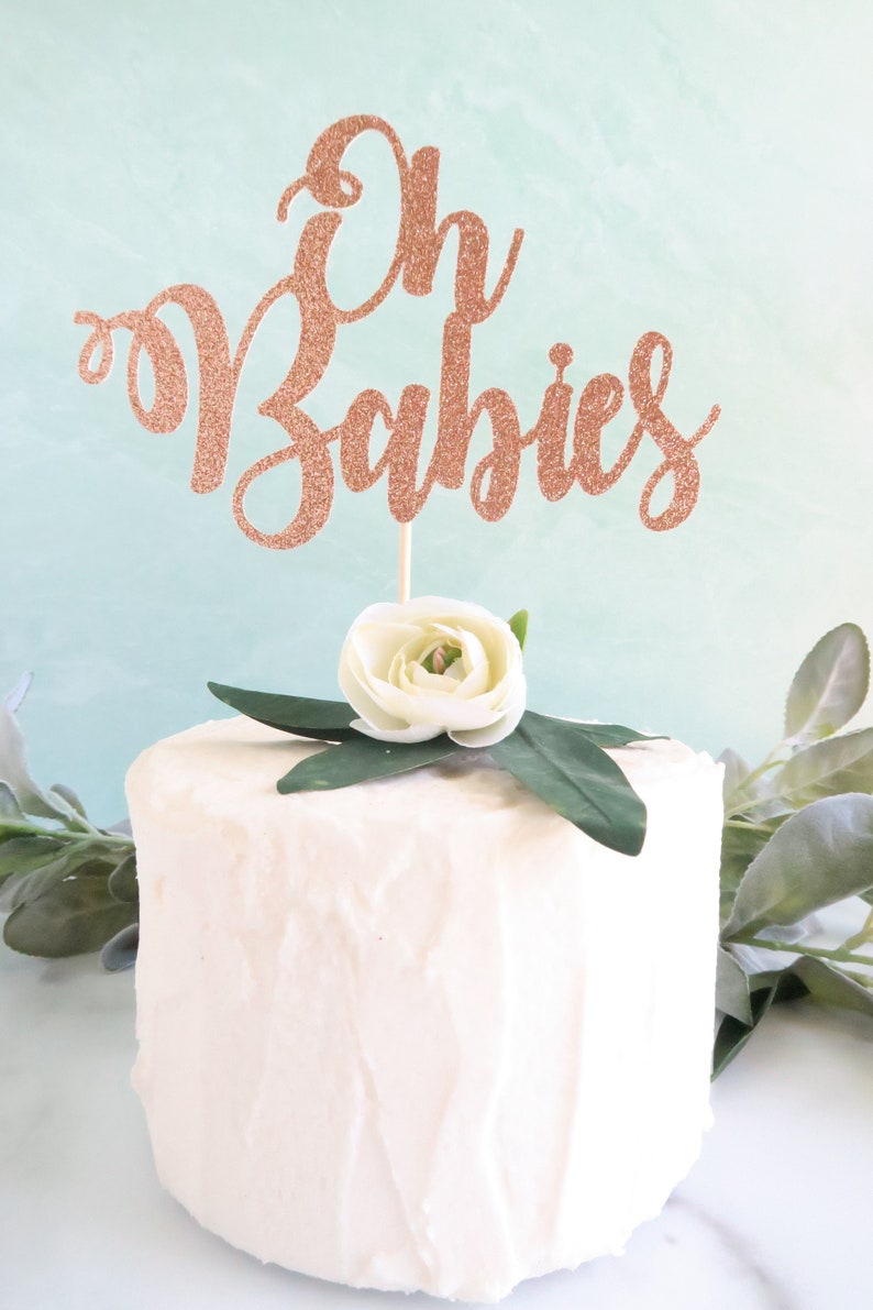 May include: A white cake with a rose gold glitter cake topper that says "Oh Babies". The cake is decorated with a white flower and green leaves.