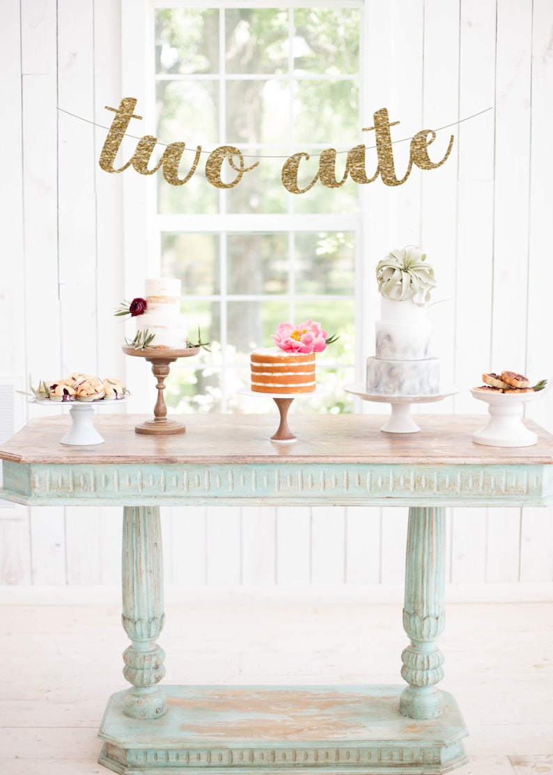May include: A light blue table with a wooden top, decorated with a gold glitter banner that reads "two cute". There are four cake stands with cakes on them, two with white frosting and two with naked cake.