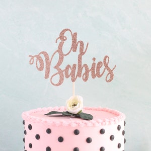 May include: A pink cake with black polka dots topped with a rose gold glitter cake topper that says "Oh Babies".