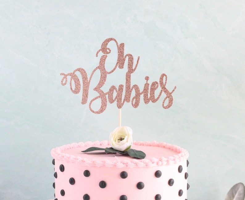May include: A pink cake with black polka dots topped with a rose gold glitter cake topper that says "Oh Babies".