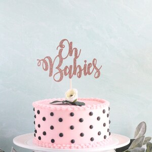 May include: Pink cake with black polka dots and a rose gold glitter cake topper that says "Oh Babies".