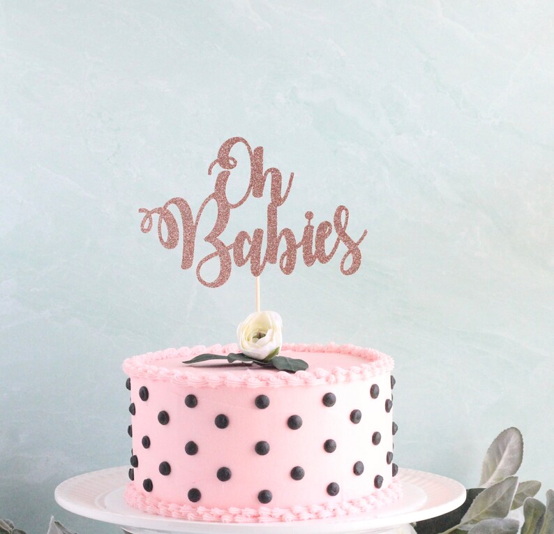 May include: Pink cake with black polka dots and a rose gold glitter cake topper that says "Oh Babies".