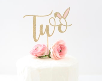 Puede incluir: Topper de pastel de purpurina dorada con la palabra "dos" y orejas de conejo rosas. El topper está en un pastel blanco con flores rosas.