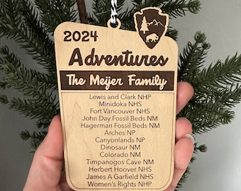 May include: Wooden Christmas ornament with the text "2024 Adventures The Meijer Family" and a list of National Park sites. The ornament has a park service logo and is hanging from a black and white string.