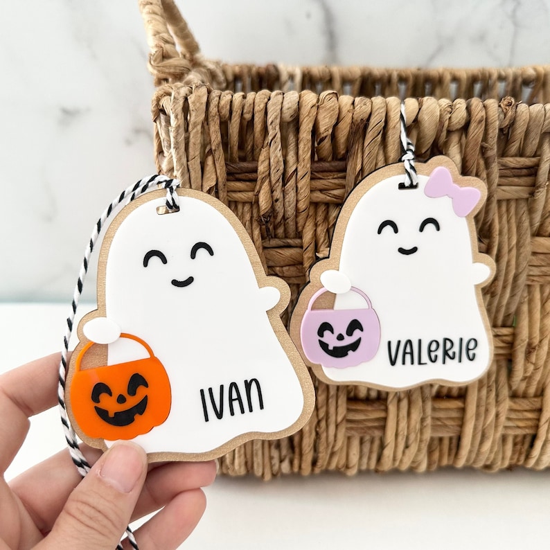 May include: Two white ghost-shaped Halloween treat bags with black eyes and smiles. One bag has a pink pumpkin and the name "Valerie" written on it. The other bag has an orange pumpkin and the name "Ivan" written on it.
