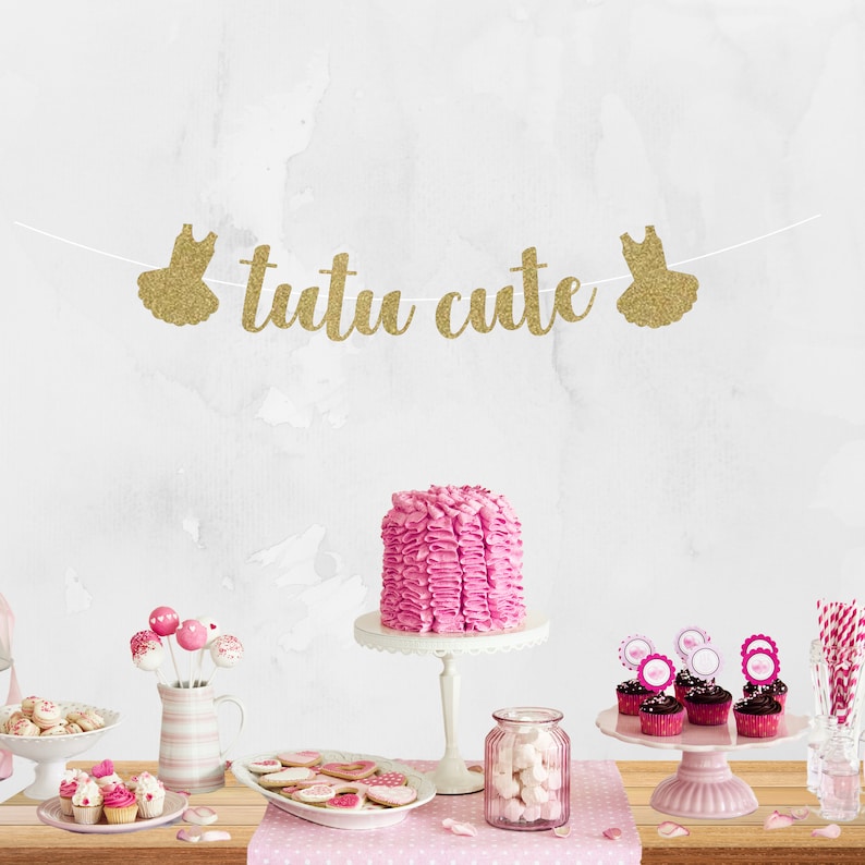May include: A pink and gold tutu cute banner with gold glitter letters and two gold glitter tutu silhouettes. The banner is hanging above a table with pink and white cupcakes, pink cake pops, a pink cake, and other pink and white decorations.