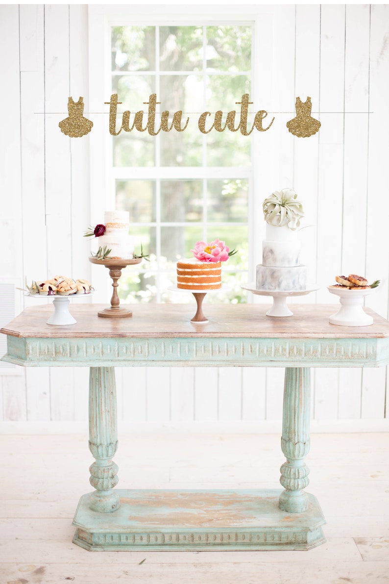 May include: A light blue table with a wood top and a gold glitter banner that says "tutu cute". There are three cakes on cake stands on the table. The cakes are decorated with flowers and greenery.