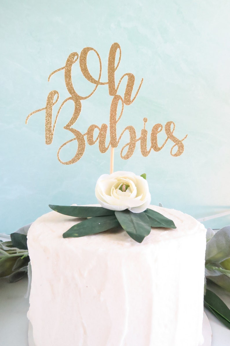Op de afbeelding: Een witte cake met gouden glitterletters die "Oh Babies" zeggen op de bovenkant. De cake is versierd met een witte bloem en groene bladeren.