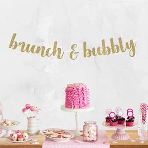 May include: A pink and gold party table setting with a cake, cupcakes, and a banner that reads "brunch & bubbly".