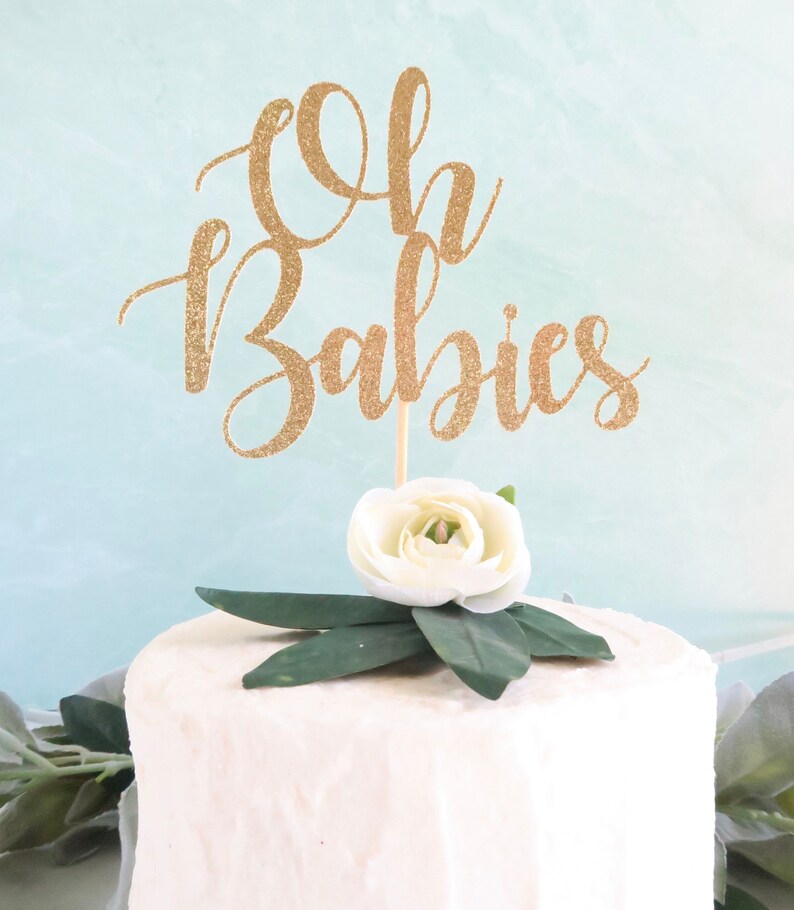 Op de afbeelding: Een gouden glittertaarttopper met de tekst "Oh Babies" en een witte bloem met groene bladeren.
