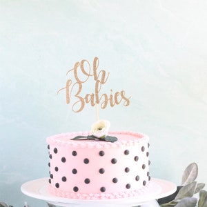 Op de afbeelding: Roze cake met zwarte stippen en een gouden glitter caketopper met de tekst "Oh Babies".