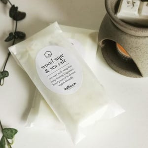 May include: Two white wax melt snap bars in clear plastic packaging. The label reads "wood sage & sea salt" and includes information about the product, such as "soy wax melt snap bar", "long-lasting fragrance", "hand poured in Devon", "vegan friendly", and the brand name "adhock".