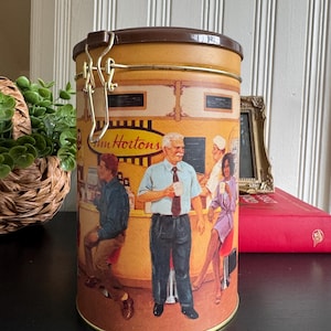 Pode incluir: Um recipiente de armazenamento cilíndrico de metal da Tim Hortons com tampa castanha articulada e fecho de metal. O exterior apresenta uma ilustração colorida de pessoas dentro de uma cafetaria, incluindo um homem de pé e outros sentados.