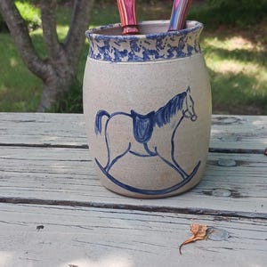 May include: A beige ceramic crock with a blue speckled rim and a blue line drawing of a rocking horse. Two multi-colored laminated wooden kitchen utensils, one solid spoon and one slotted spoon, are placed inside the ceramic container.