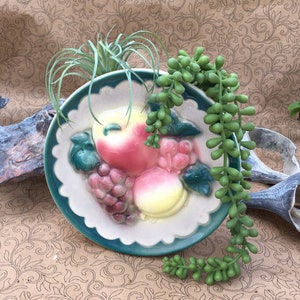 May include: A vintage ceramic wall planter with a green and white design featuring a raised image of fruit. The planter is decorated with a scalloped edge and is filled with green succulents.