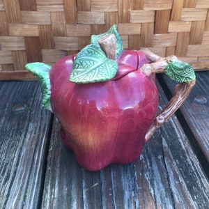 May include: A red ceramic teapot shaped like an apple with green leaves and a brown twig handle.