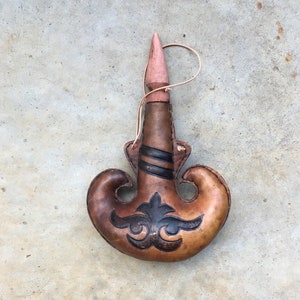May include: A brown leather flask with a wooden stopper and a decorative design. The flask has a strap for carrying.