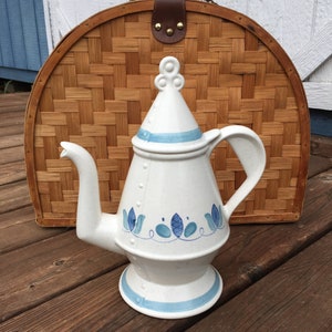 May include: A white ceramic teapot with a blue and white floral design. The teapot has a round lid with a knob on top and a spout on the side. The teapot is sitting on a wooden surface.