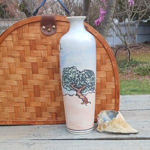 May include: A white ceramic vase with a hand-painted design of a tree with a pink and blue sky. The vase is sitting on a wooden table.