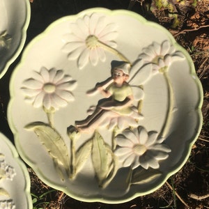 May include: A white ceramic plate with a raised design of a fairy sitting on a daisy. The plate is decorated with green leaves and pink and white daisies.