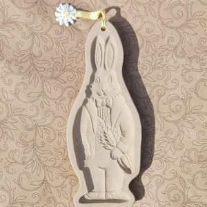 May include: A white ceramic cookie cutter shaped like a rabbit wearing a suit and holding a bouquet of flowers. The cutter has a loop at the top for hanging.