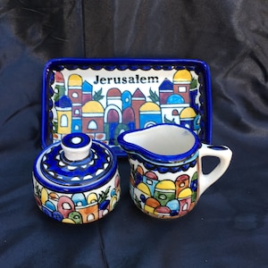 May include: A blue and white ceramic tray with a Jerusalem cityscape design. The tray is accompanied by a matching creamer and sugar bowl, both featuring the same colorful cityscape design.
