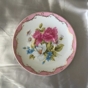 May include: A white porcelain saucer with a pink and gold rim. The center features a floral design with pink, white, and blue flowers.