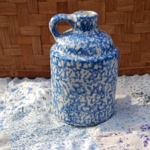 May include: A blue and white ceramic jug with a handle. The jug has a speckled pattern and a rounded body. The background includes a woven basket and a floral quilt.
