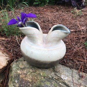 May include: A white ceramic vase with a brown glaze and two spouts. The vase is sitting on a rock and has a purple flower inside.