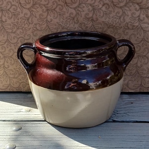 May include: A brown and white glazed ceramic crock with two handles. The crock is sitting on a wooden surface.